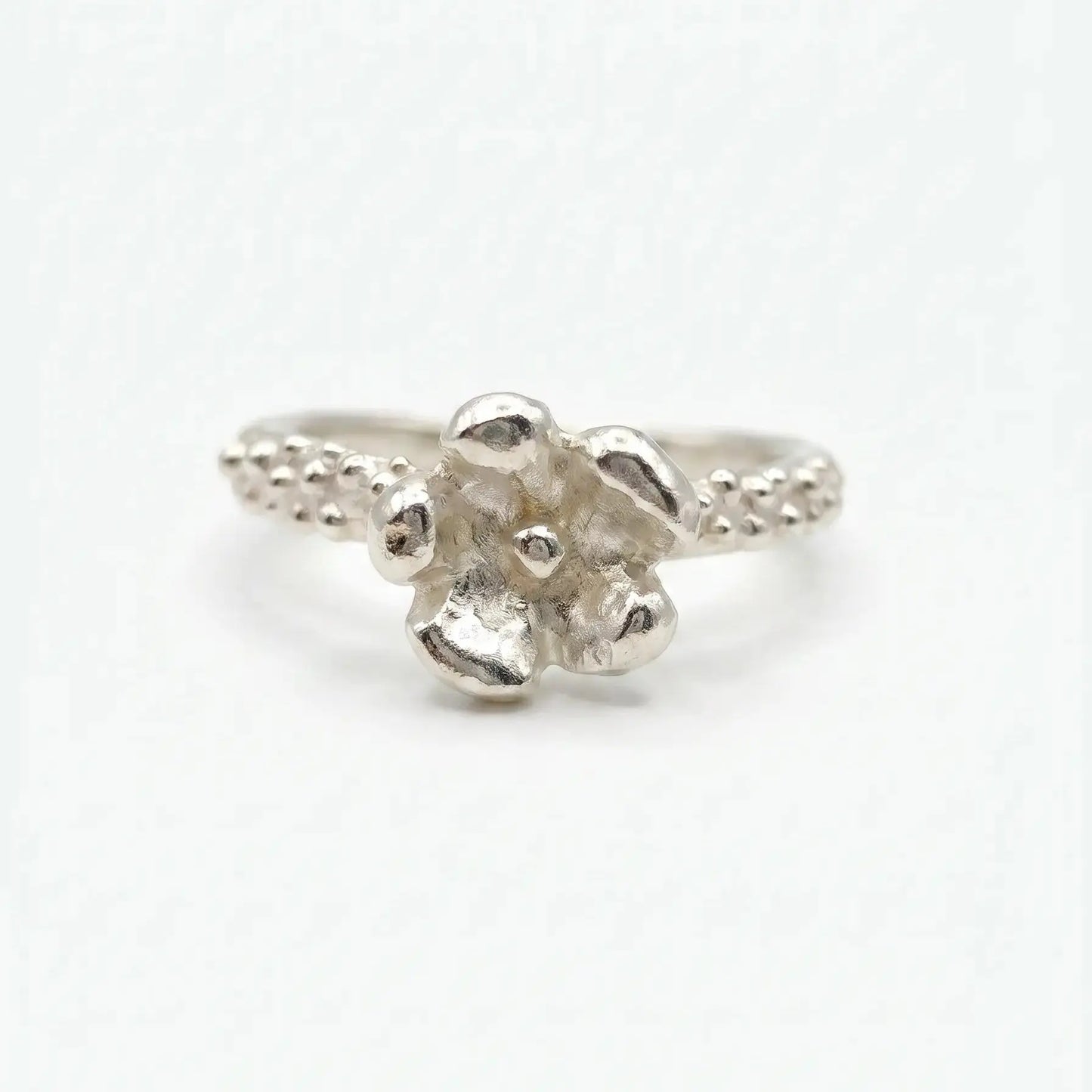 Anello artigianale in argento con gambo sottile caratterizzato da una texture puntinata e un dettaglio a forma di piccolo fiore a cinque petali sulla sommità. Lo stile è organico e materico, presentato su uno sfondo bianco neutro in posizione frontale