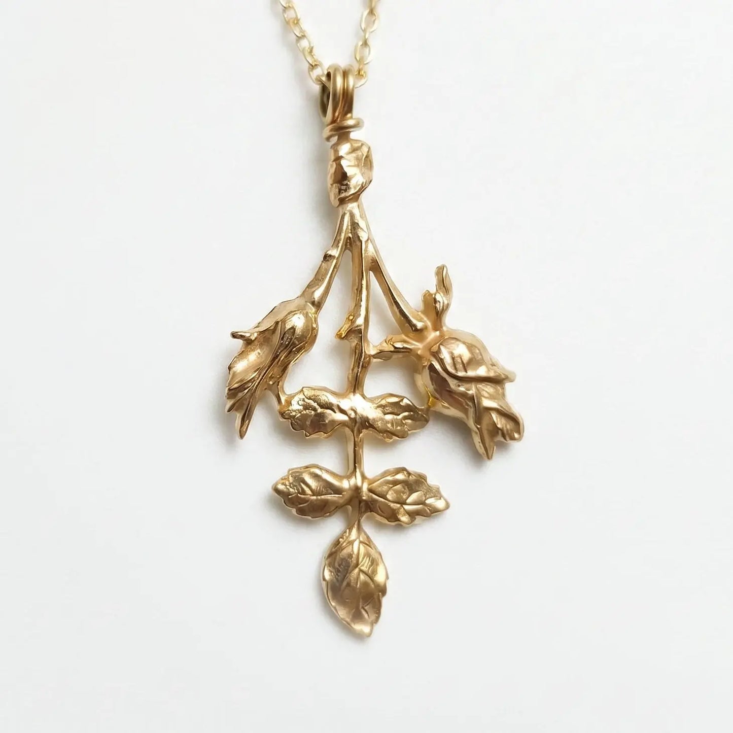 Un primo piano di una collana sottile in oro con un pendente botanico che raffigura due boccioli di rosa e un rametto con foglie finemente dettagliate. Il gioiello, adagiato su uno sfondo bianco pulito, mostra una texture materica e scultorea che riflette la luce