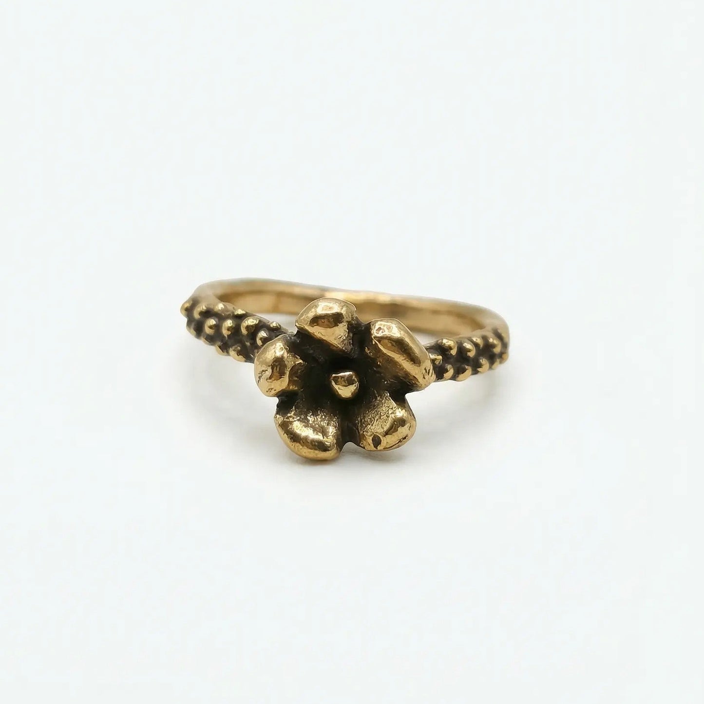 Anello in bronzo dorato fatto a mano, design unico con fiore a cinque petali e gambo testurizzato a granulazione con brunitura negli incavi, su sfondo bianco.