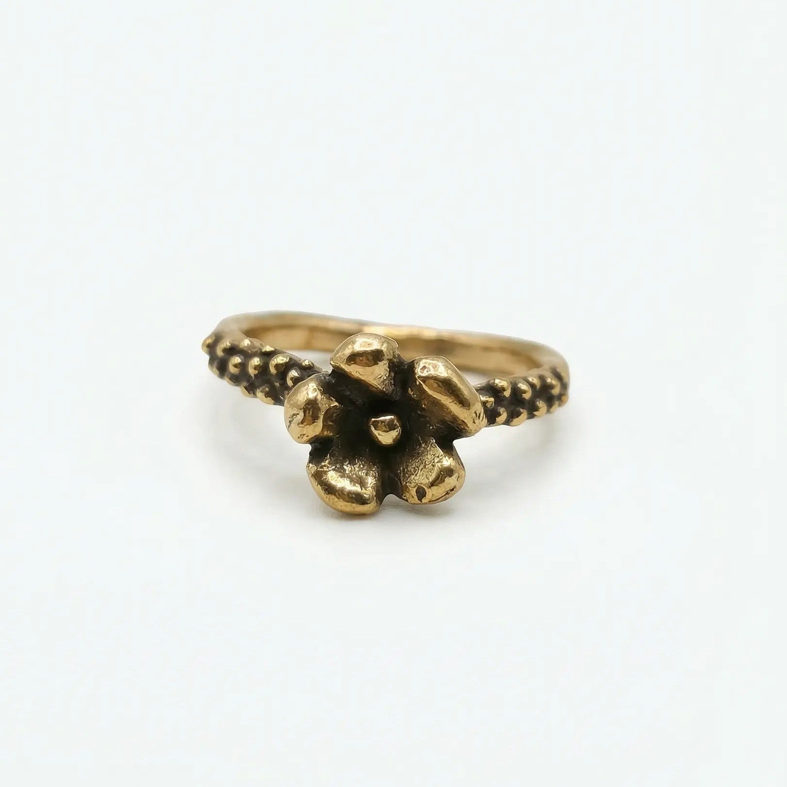 Anello in bronzo dorato fatto a mano, design unico con fiore a cinque petali e gambo testurizzato a granulazione con brunitura negli incavi, su sfondo bianco.