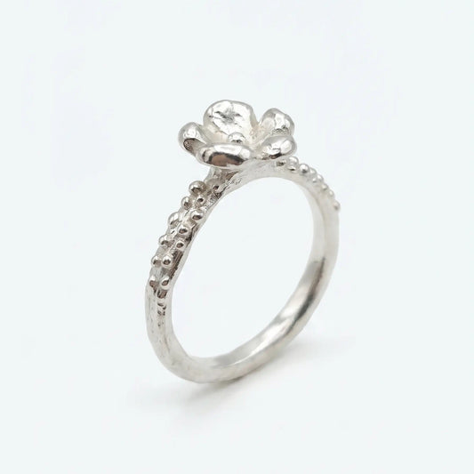 Anello artigianale in argento con gambo sottile caratterizzato da una texture puntinata e un dettaglio a forma di piccolo fiore a cinque petali sulla sommità. Lo stile è organico e materico, presentato su uno sfondo bianco neutro