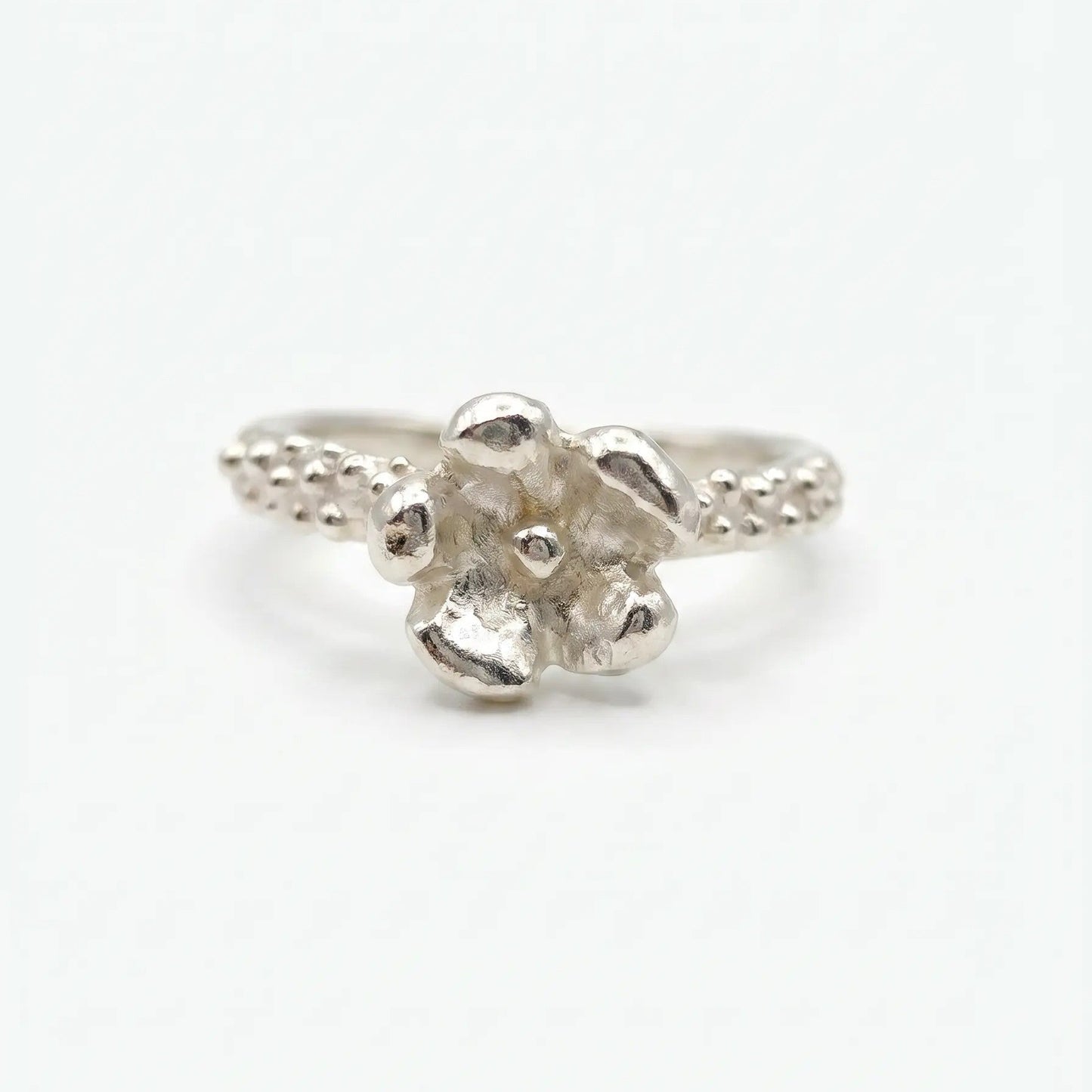 Anello artigianale in argento con gambo sottile caratterizzato da una texture puntinata e un dettaglio a forma di piccolo fiore a cinque petali sulla sommità. Lo stile è organico e materico, presentato su uno sfondo bianco neutro in posizione frontale