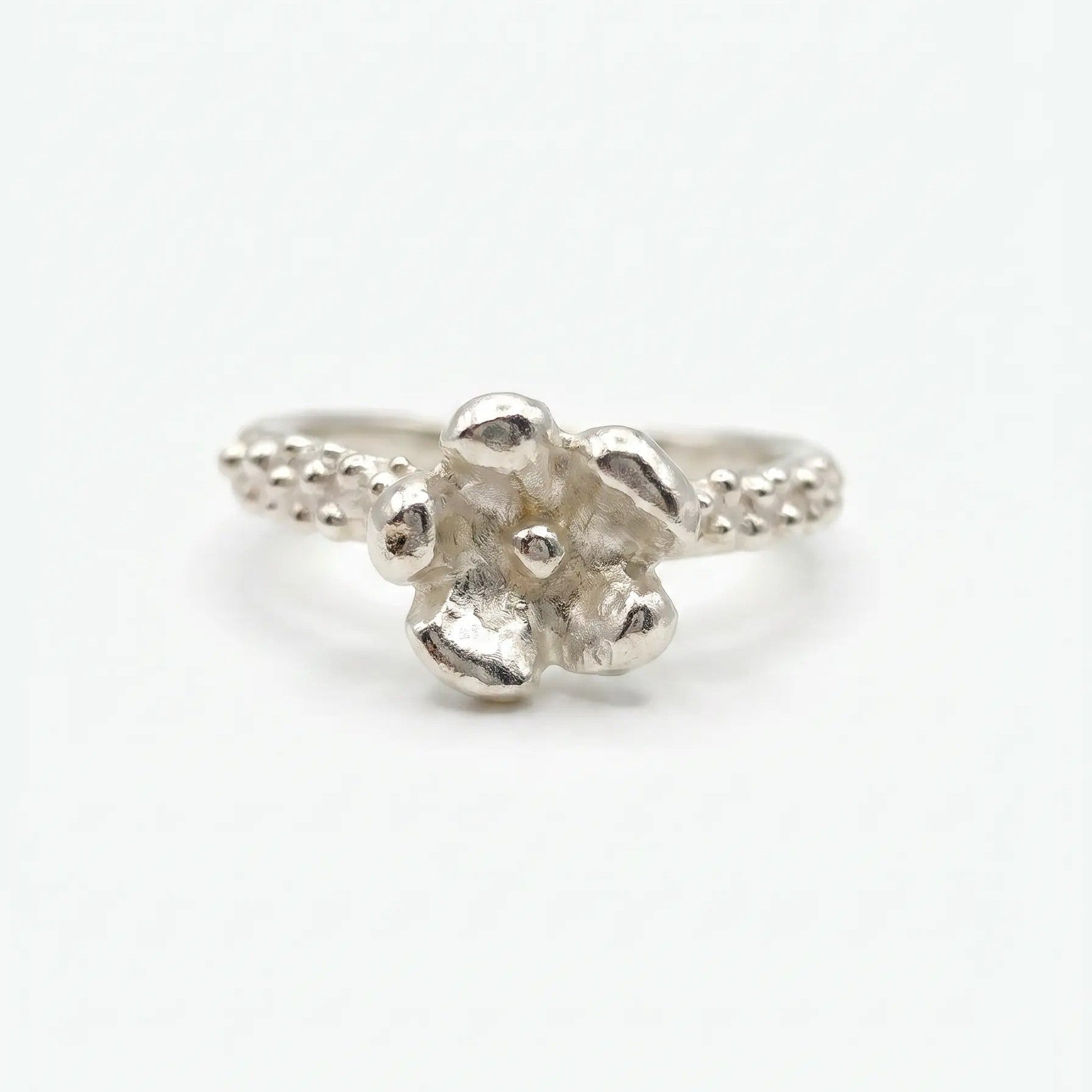 Anello artigianale in argento con gambo sottile caratterizzato da una texture puntinata e un dettaglio a forma di piccolo fiore a cinque petali sulla sommità. Lo stile è organico e materico, presentato su uno sfondo bianco neutro in posizione frontale
