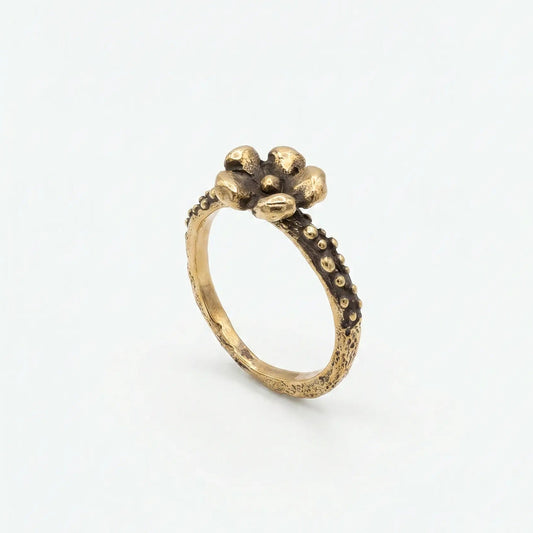Anello in bronzo dorato fatto a mano con fiore stilizzato a cinque petali e gambo a granulazione con brunitura, su sfondo bianco neutro.