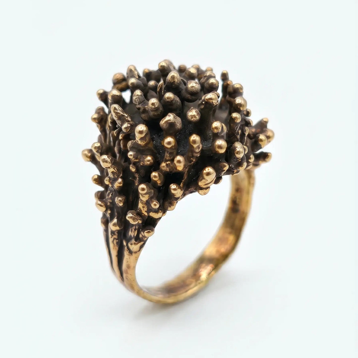 Anello artigianale in bronzo con design organico a rilievo, dettaglio di texture rugosa con punte arrotondate dorate, vista di tre quarti su sfondo bianco.