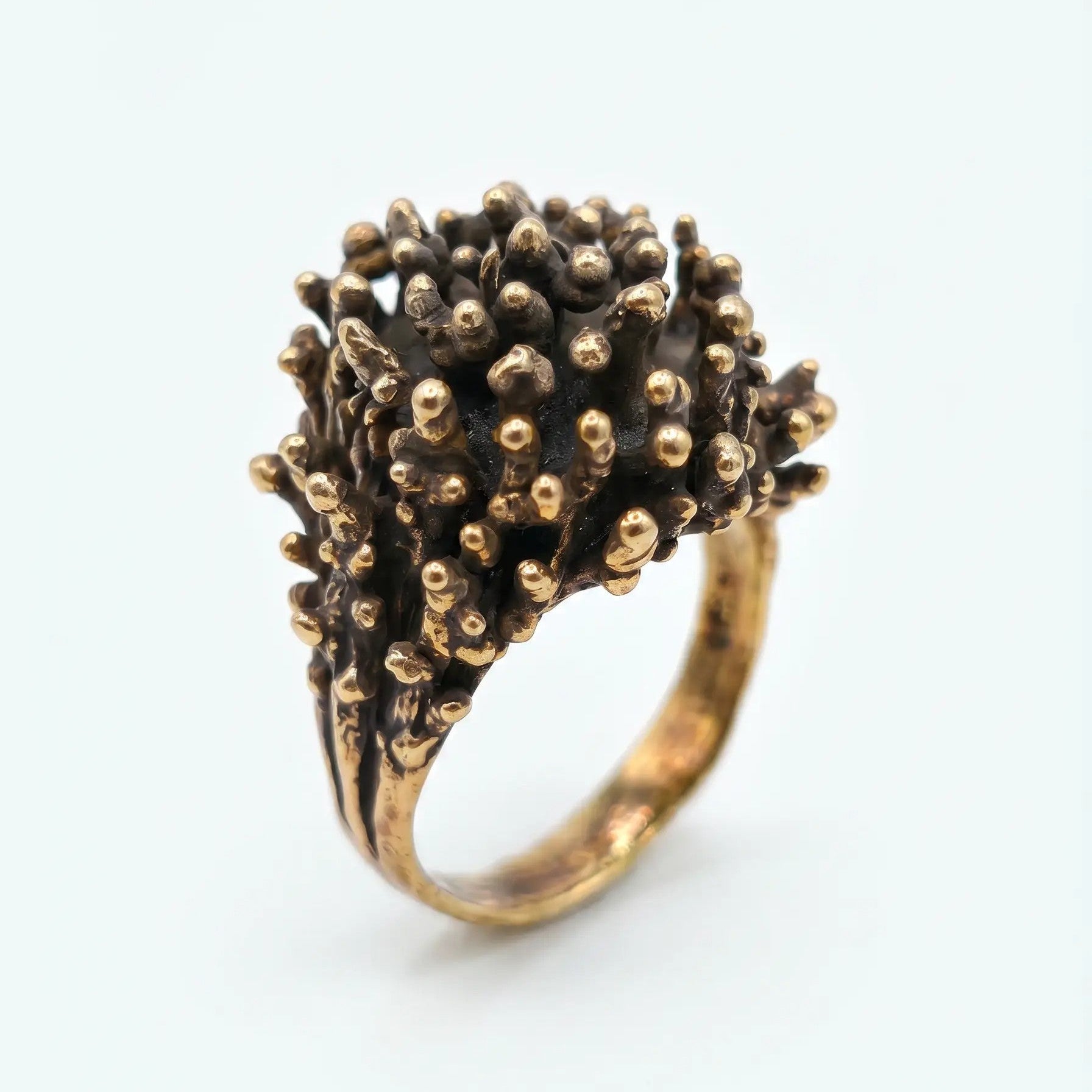 Anello artigianale in bronzo con design organico a rilievo, dettaglio di texture rugosa con punte arrotondate dorate, vista di tre quarti su sfondo bianco.