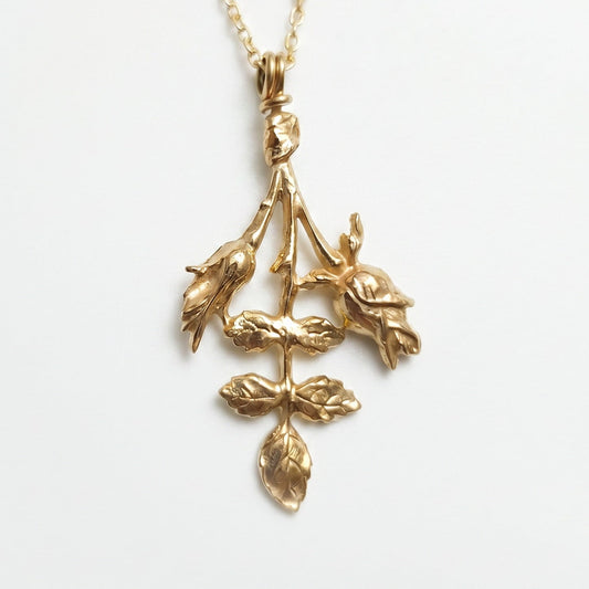 Un primo piano di una collana sottile in oro con un pendente botanico che raffigura due boccioli di rosa e un rametto con foglie finemente dettagliate. Il gioiello, adagiato su uno sfondo bianco pulito, mostra una texture materica e scultorea che riflette la luce